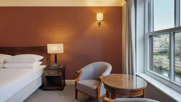 A hotel room with a bed, a chair, a small table, a nightstand, a lamp, and a window with a view of a river.