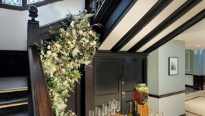 Una escalera decorada con arreglos florales, una mesa con vasos y botellas, y una puerta con un cuadro enmarcado.