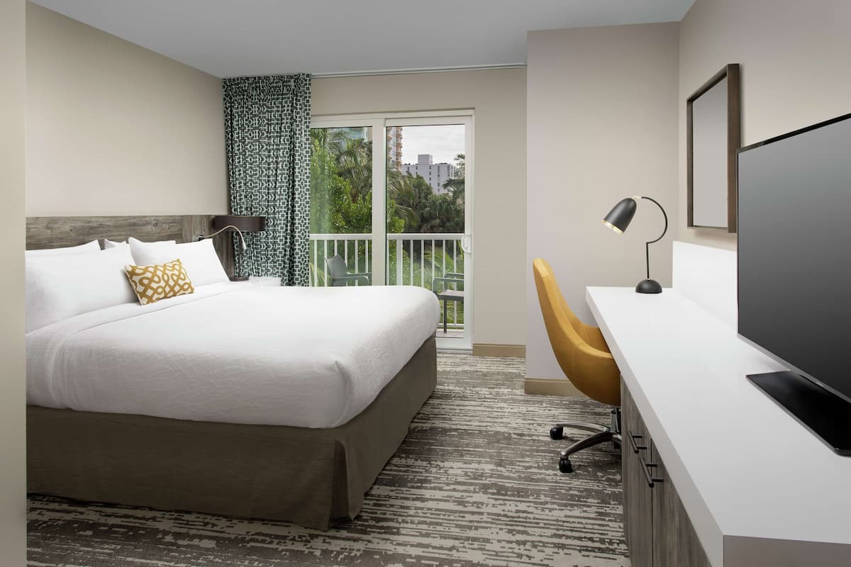 A hotel room with a bed, a desk, a television, and a balcony with a view of palm trees and buildings.