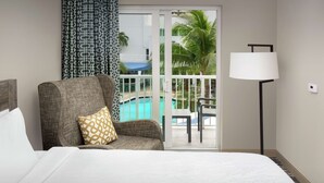 Une chambre d’hôtel avec un lit, un fauteuil gris orné d’un coussin à motifs, une lampe sur pied et une vue sur une piscine et des palmiers, visible à travers une porte vitrée.