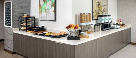 A modern kitchen counter with a variety of food items, including fruits, pastries, and beverages, along with a chalkboard menu and decorative artwork.