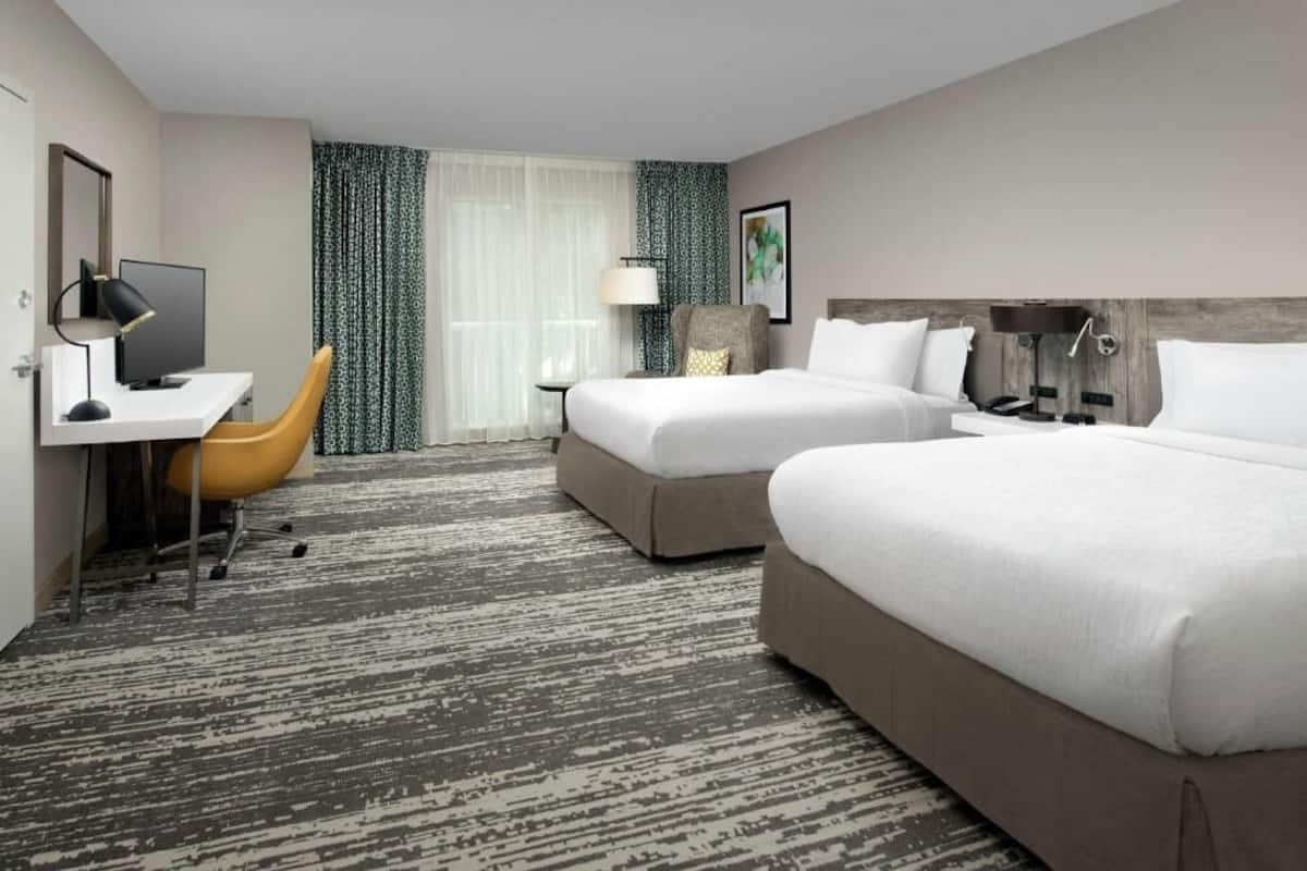 A hotel room with two beds, a desk with a computer, a chair, a lamp, and a large window with curtains.