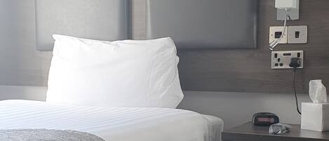 A hotel room with a large bed, a bedside table with a lamp, and a wall-mounted power outlet.