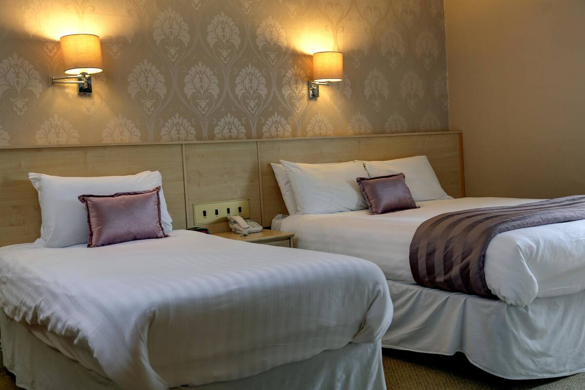 Une chambre d’hôtel avec deux lits, chacun agrémenté de draps blancs et d’un seul oreiller violet. Des lampes murales sont fixées de chaque côté des lits, et un téléphone se trouve sur la table de chevet entre les deux.