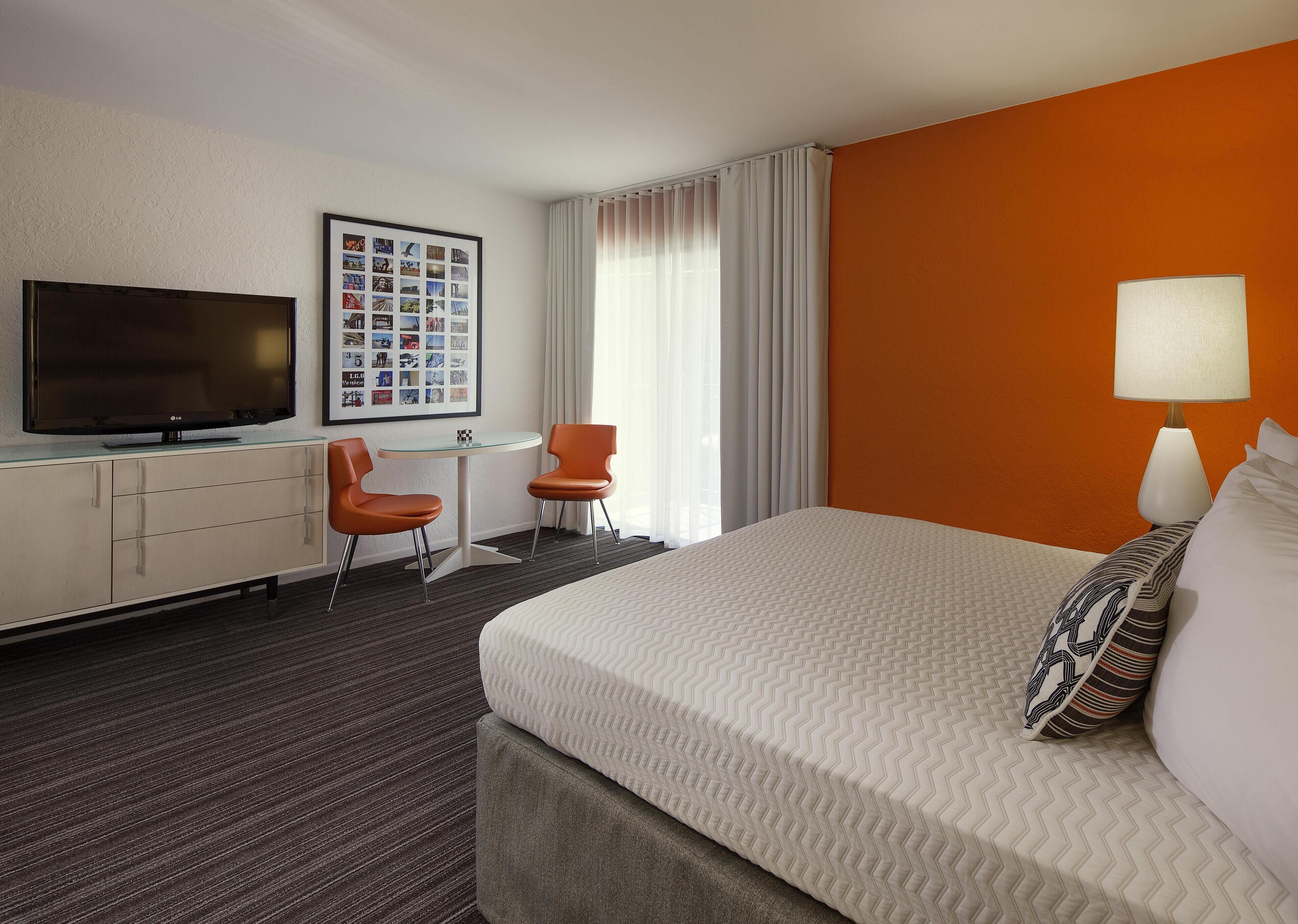 A hotel room with a bed, a desk with a TV, two chairs, a framed photo collage, and a lamp.