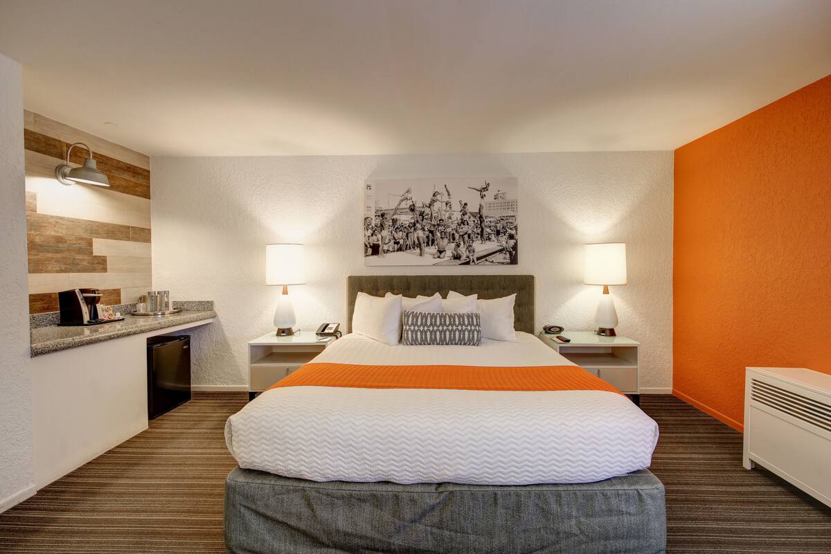 Ein Hotelzimmer mit einem Bett, zwei Nachttischen, einem Schreibtisch, einem Stuhl, einer kleinen Küchenzeile und einem Wandbild.