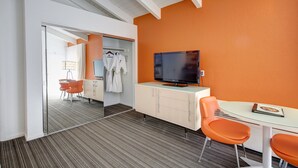 A hotel room with a glass partition, a television on a cabinet, an orange wall, and a dining area with orange chairs. - The Inn at Venice Beach (Venice)
