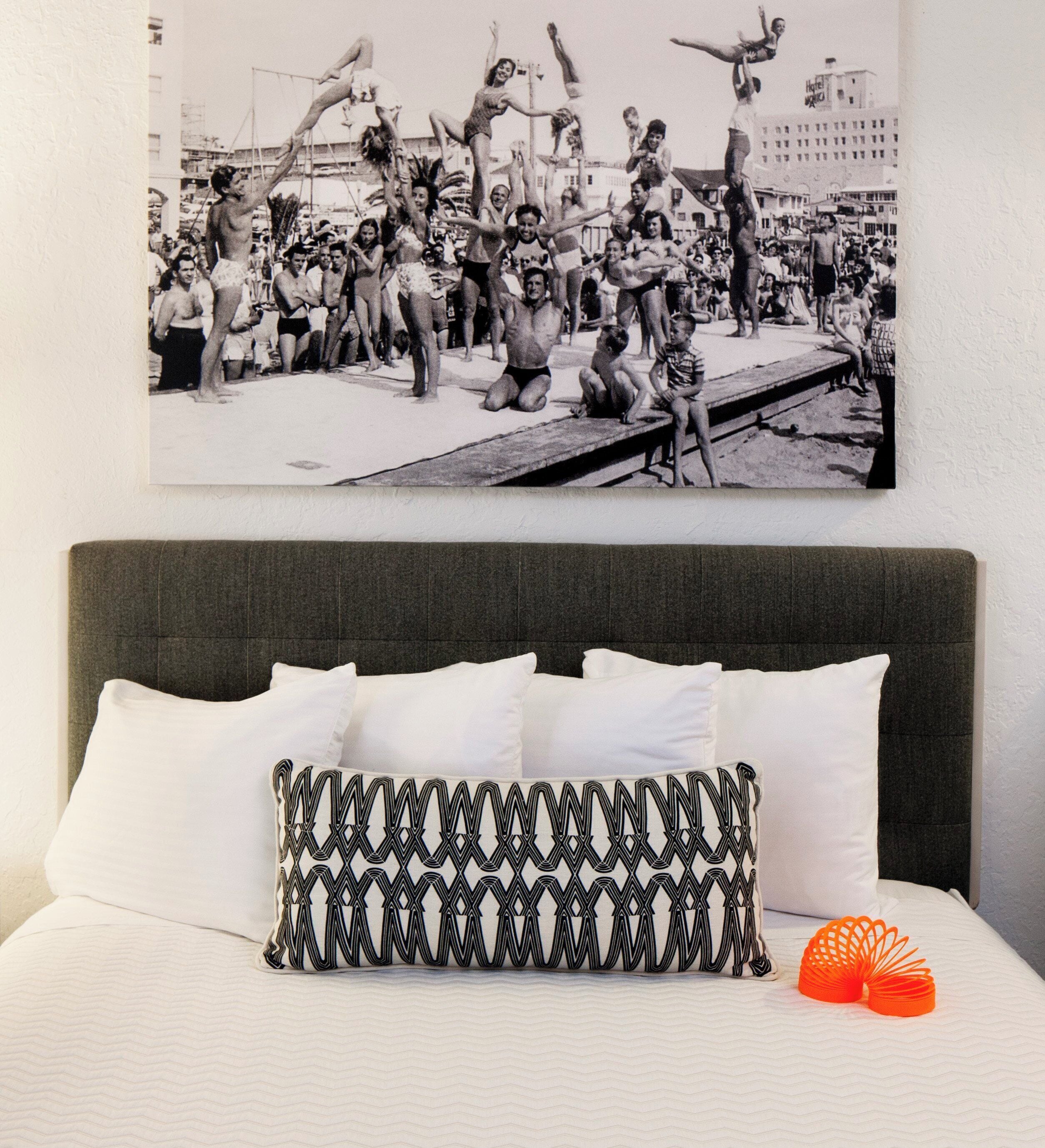 A bed with white pillows and a patterned pillow, a black and white photograph on the wall, and a small orange object on the bed.