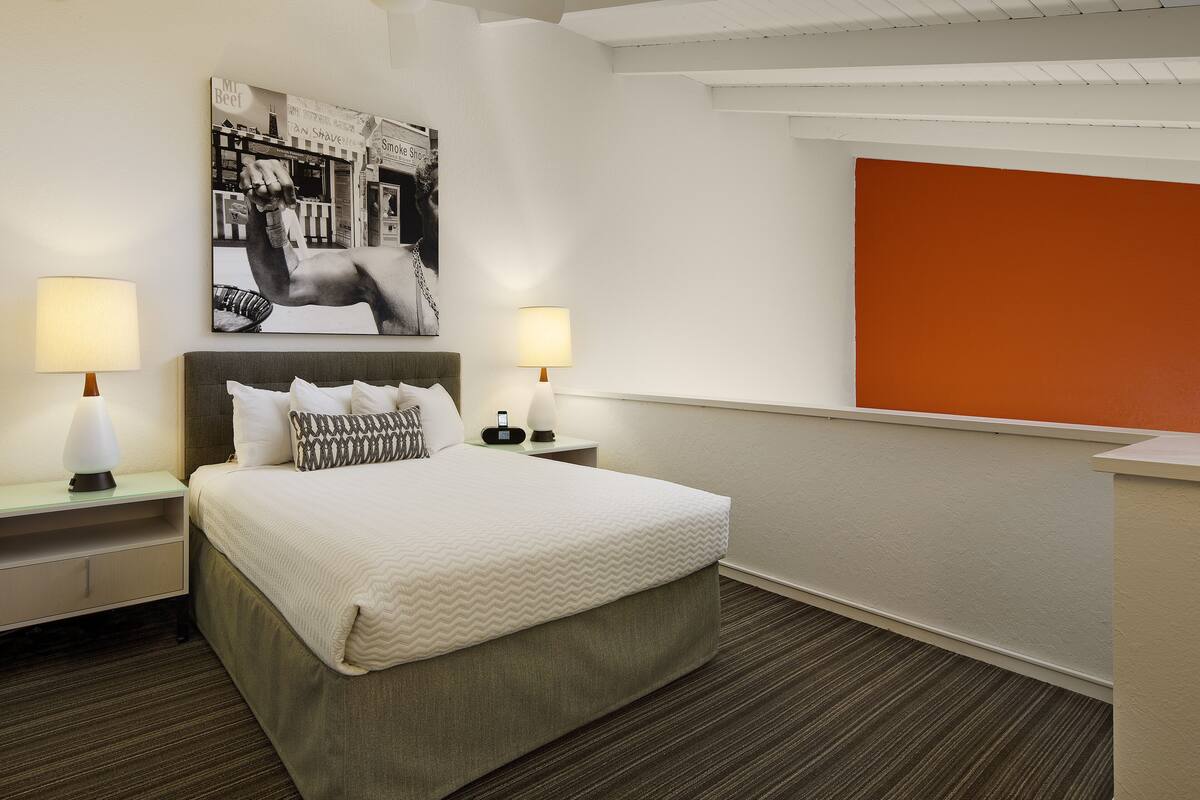 Ein Hotelzimmer mit einem Bett, zwei Nachttischen, einem Wandbild und einer leuchtend orangefarbenen Wand.