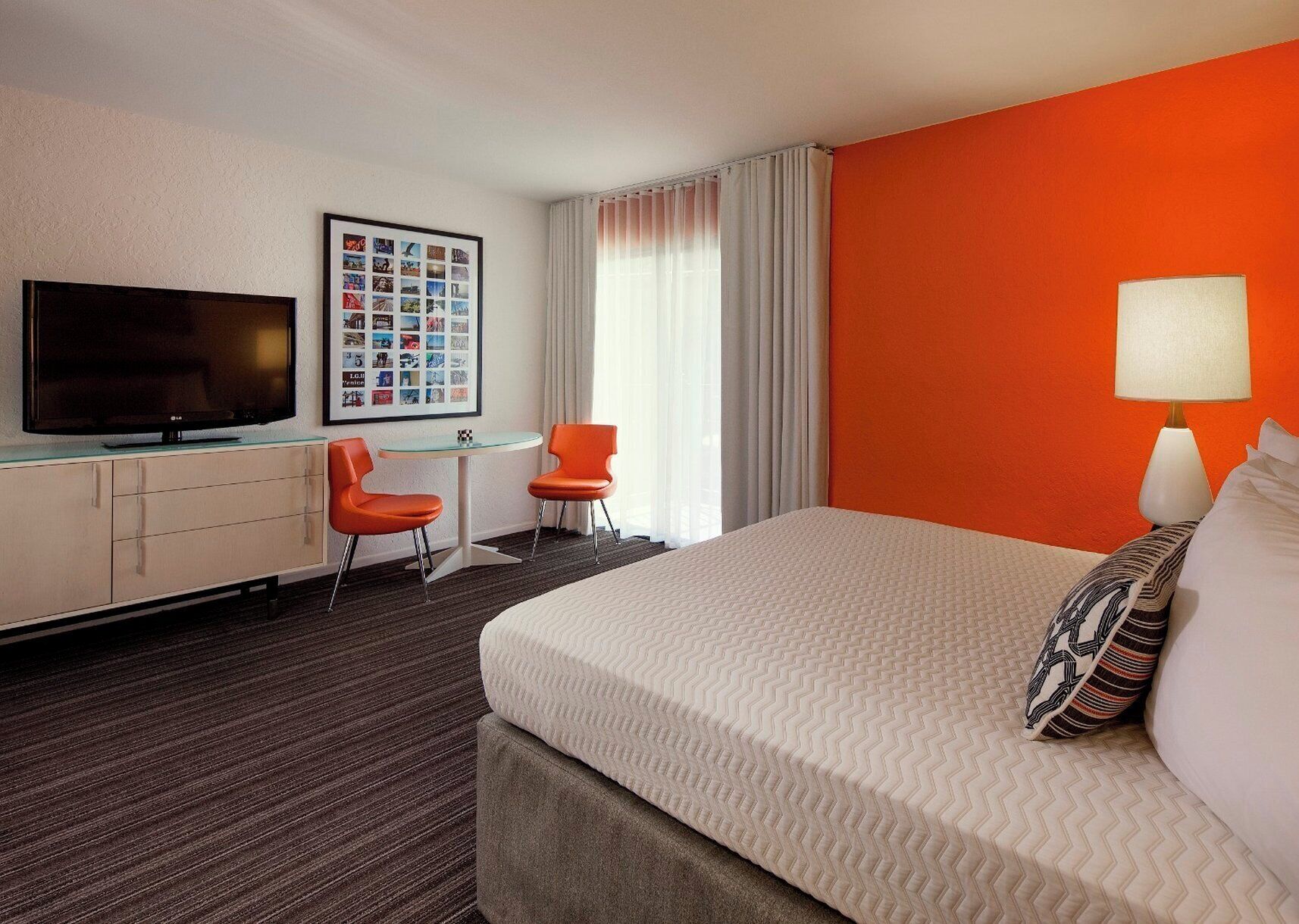 Habitación de hotel con una cama, televisión, escritorio y dos sillas, un collage de fotos enmarcado, y una ventana con cortinas.