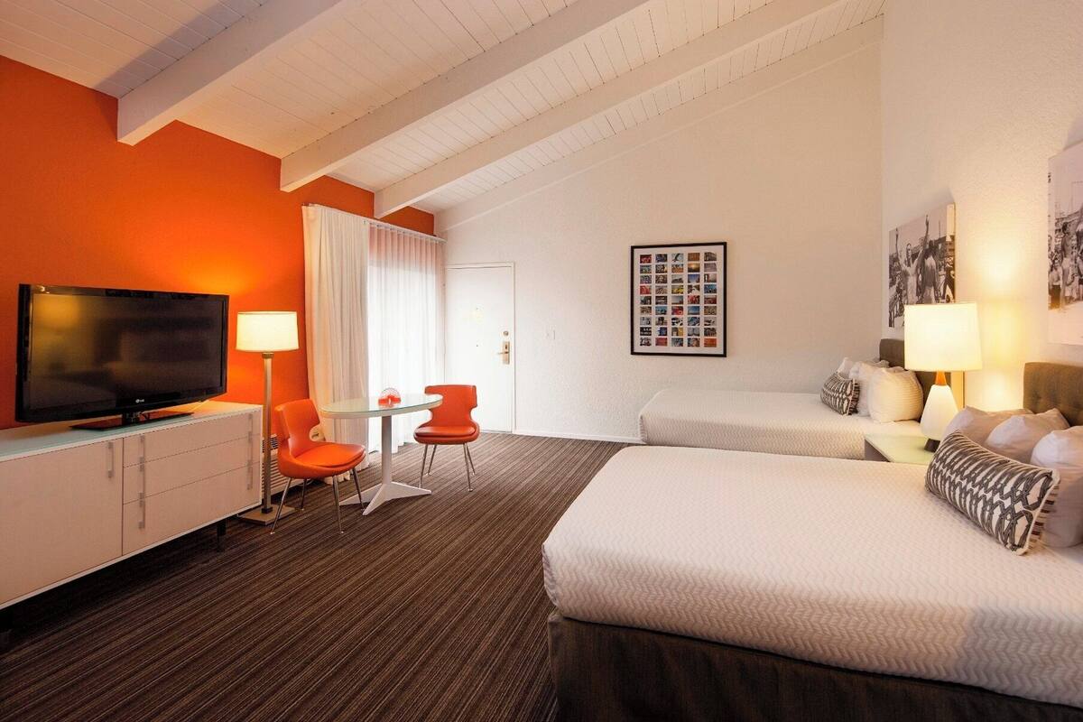 Ein Hotelzimmer mit Fernseher, zwei Betten, einem Schreibtisch mit Stuhl und einem gerahmten Foto an der Wand.
