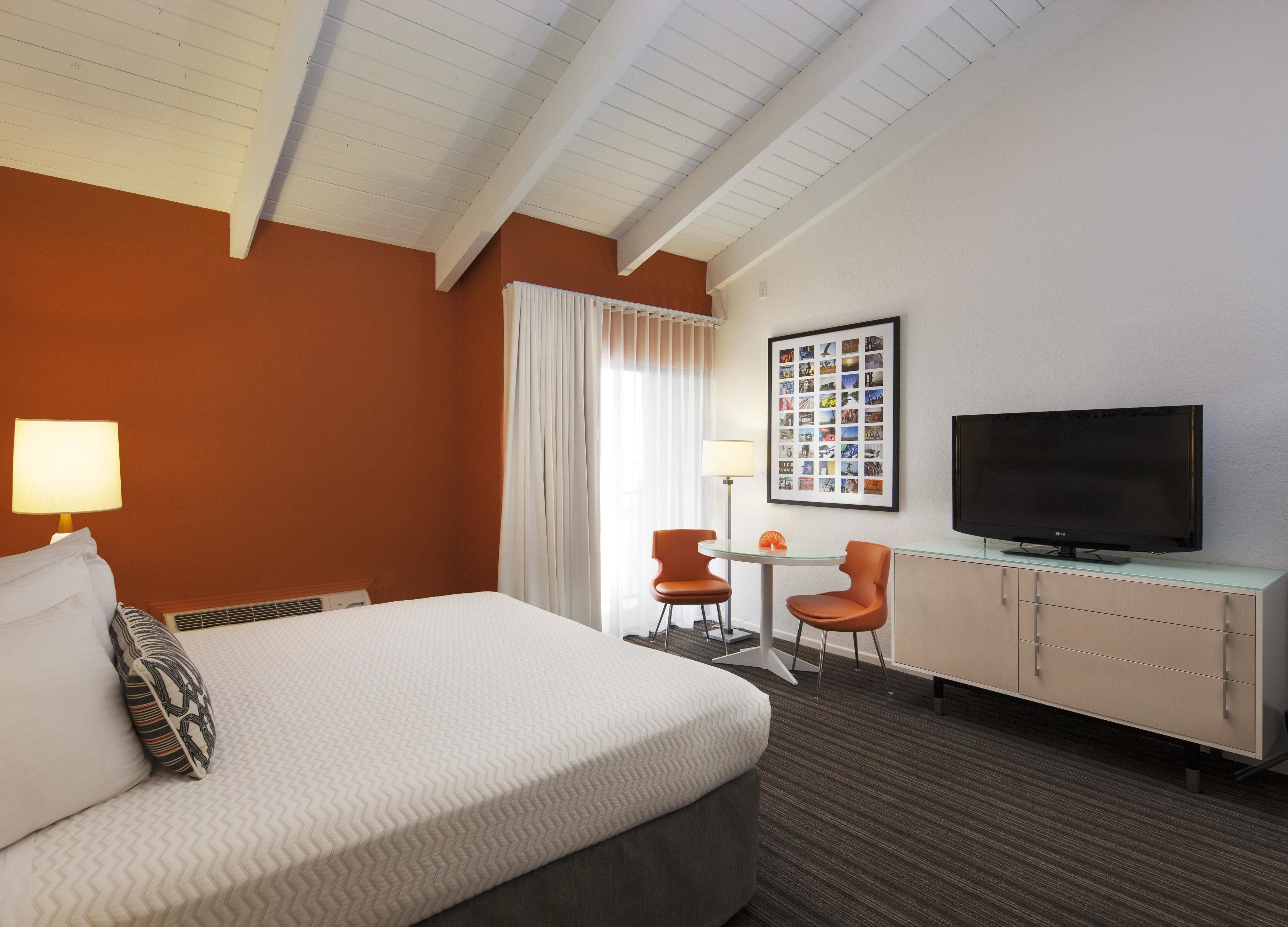 Une chambre d’hôtel avec un lit, un bureau avec une télévision, deux chaises, une petite table et un cadre photo accroché au mur.