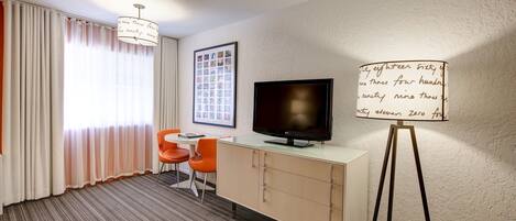 A hotel room with a television on a stand, a desk with a chair, a large window with curtains, and a wall-mounted whiteboard.