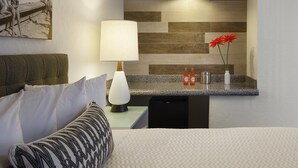 A hotel room with a bed, a bedside table with a lamp, a small kitchen area with a sink and a mirror, and a wall with a wooden panel design. - The Inn at Venice Beach (Venice)