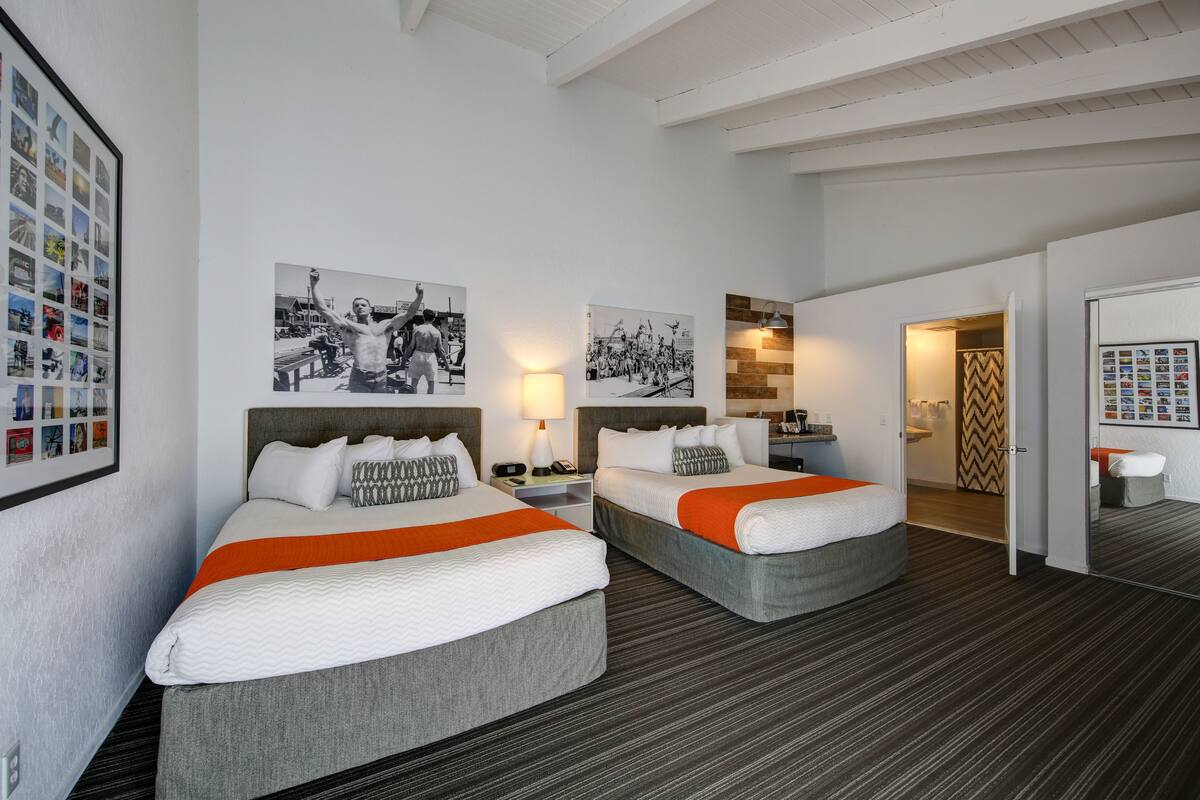 Ein Hotelzimmer mit zwei Betten, einer großen gerahmten Fotocollage an der Wand und einem durch eine offene Tür einsehbaren Badezimmer.