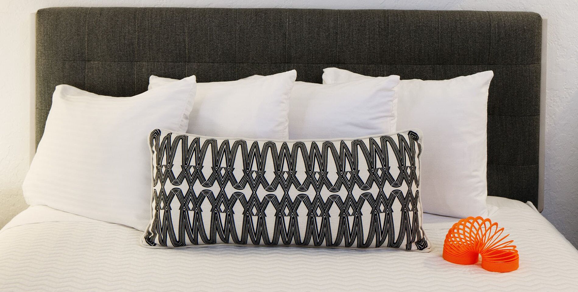 A bed with white pillows and a patterned pillow, a grey headboard, and two orange decorative pillows.