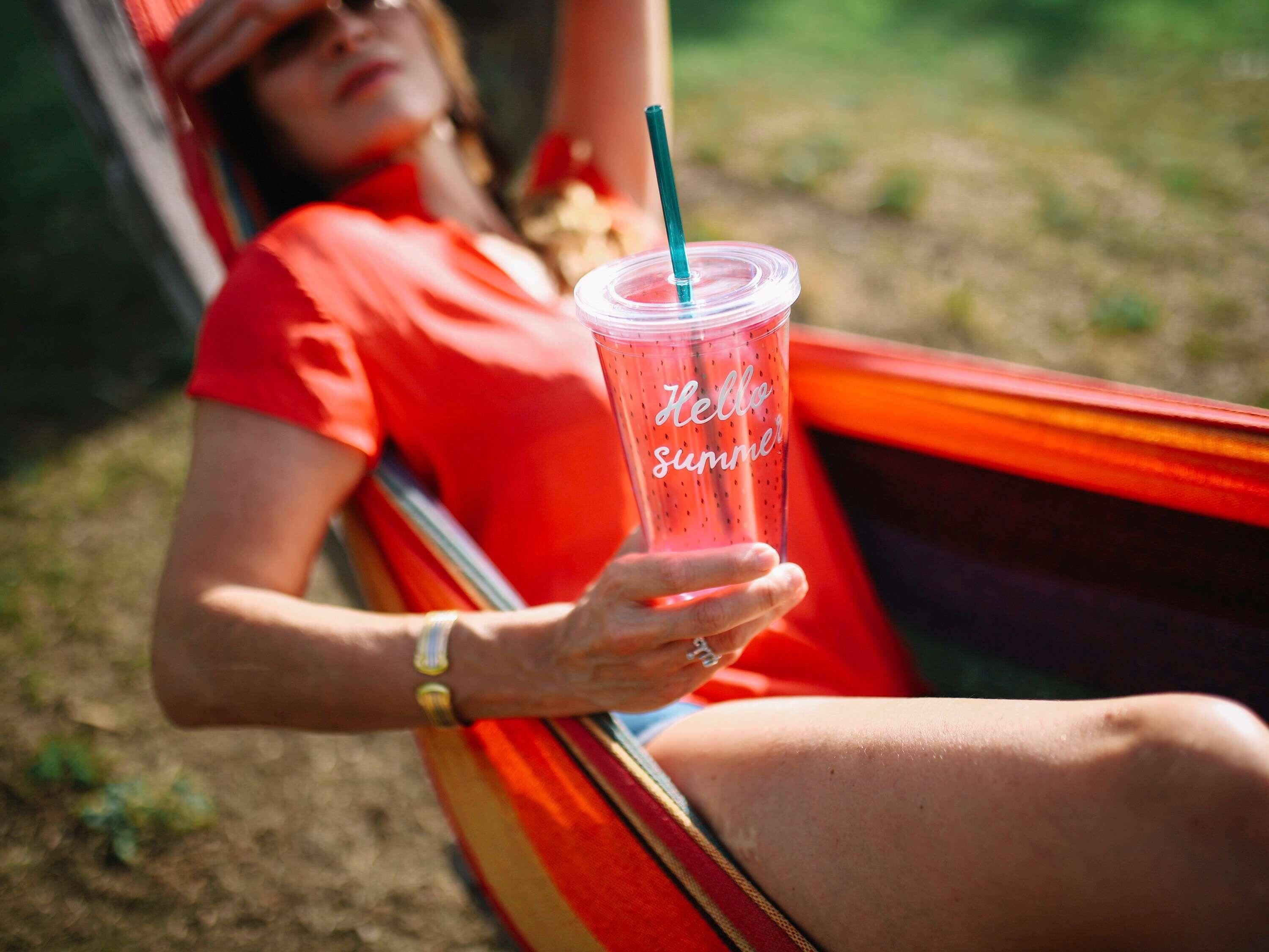 A person reclining in a hammock outdoors, holding a clear plastic cup with a straw and the words "Hello Summer" on it.