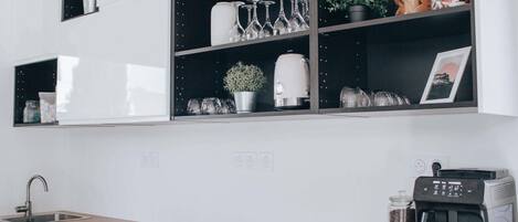 A kitchen with white cabinets, a wooden countertop, a coffee machine, and shelves with glassware and plants.