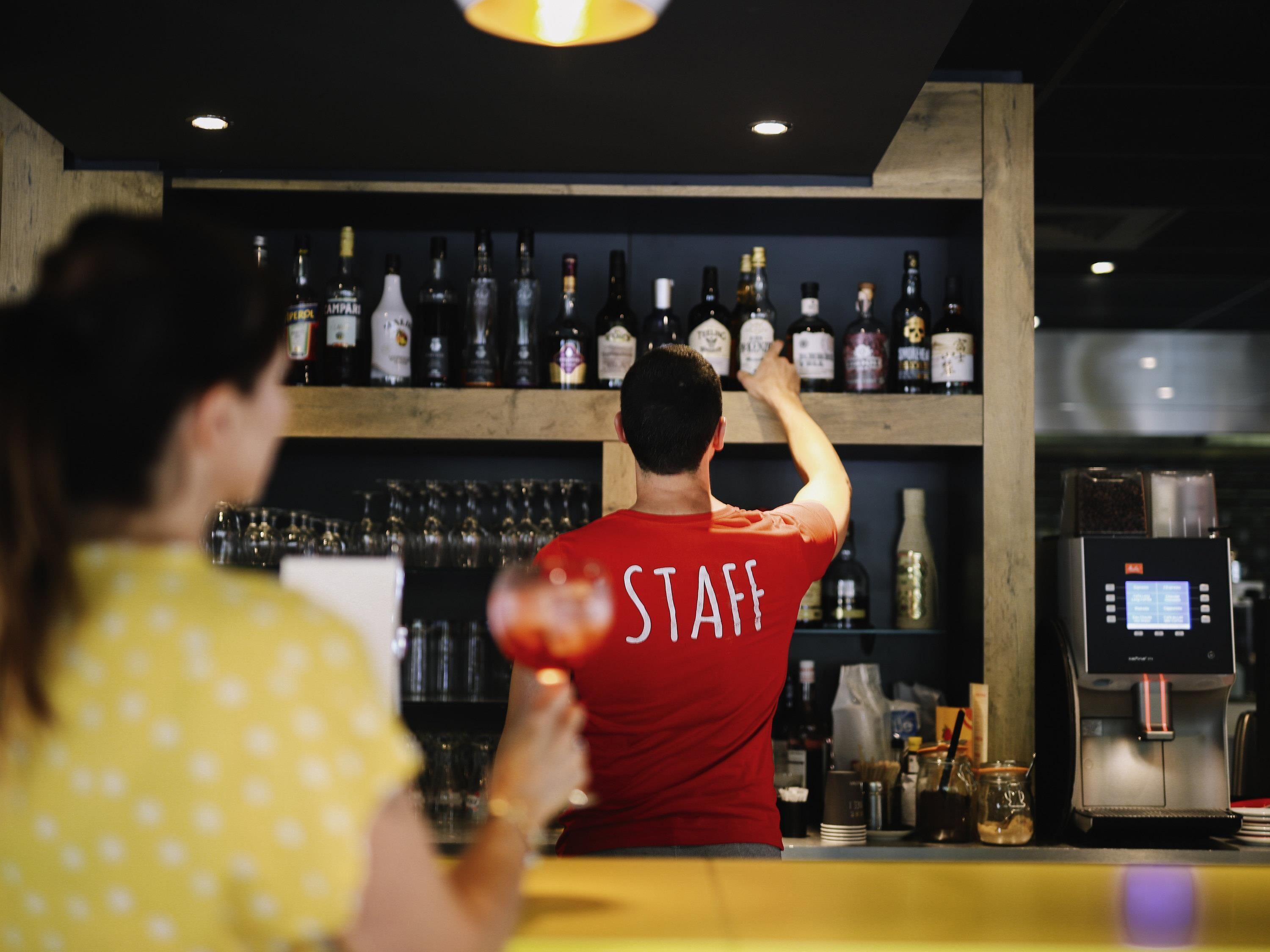 A bar area with a bartender in a red shirt labeled "STAFF" behind the counter, a woman in a yellow polka dot shirt in the foreground, and a variety of liquor bottles on shelves.