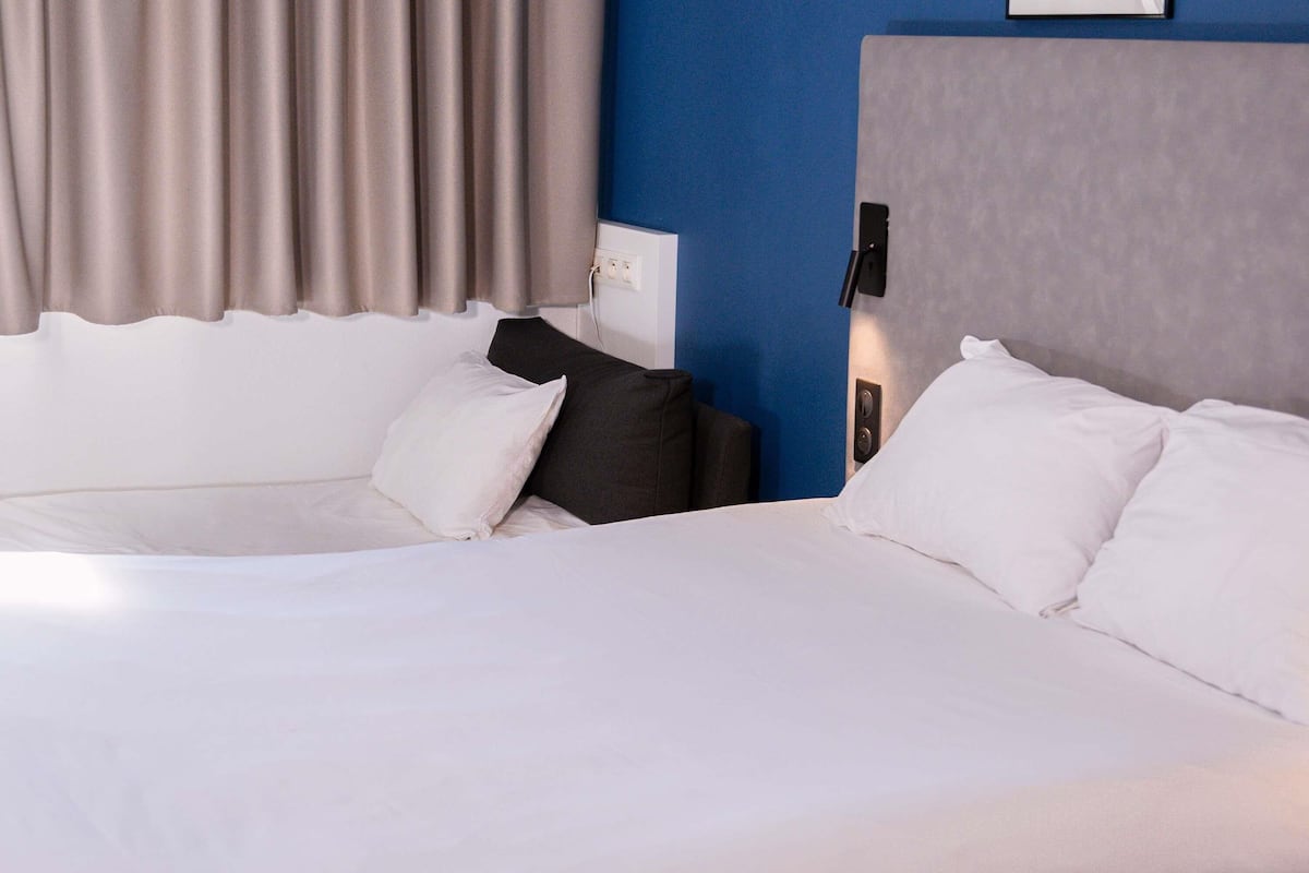 A hotel room with a bed, pillows, a headboard, a wall socket, and a curtain.