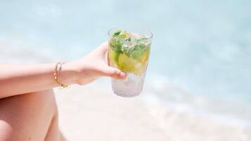 A person holding a glass of a beverage with lime and mint, at a beach with clear water.