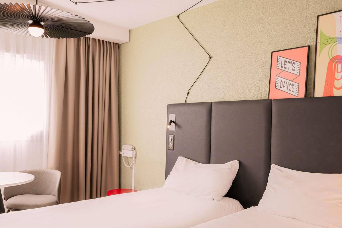 A hotel room with a bed, a chair, a small table, a phone, and a wall with a poster.