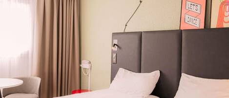 A hotel room with a bed, a chair, a small table, a phone, and a wall with a poster.