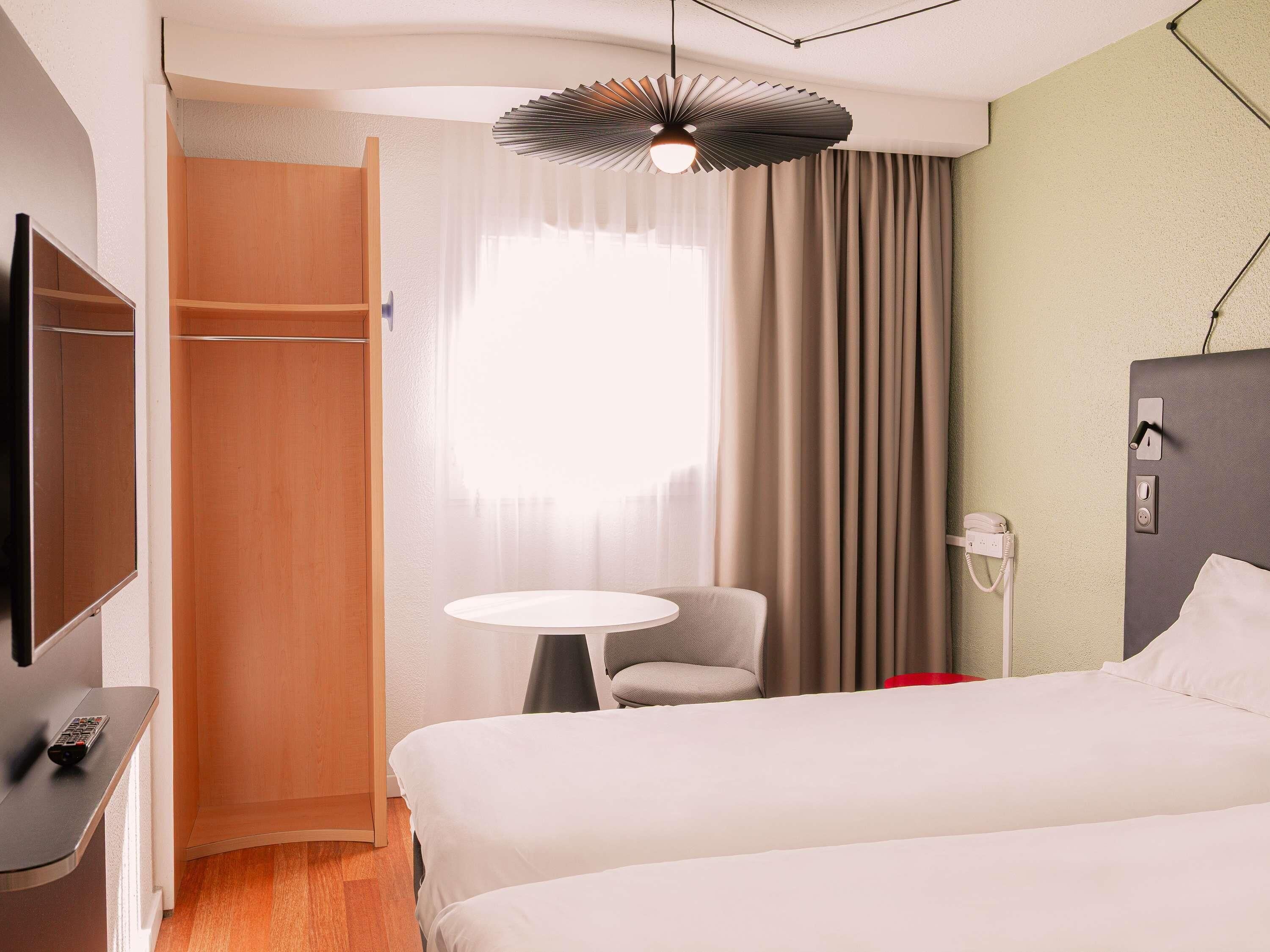 A hotel room with a bed, a desk with a TV, a chair, a small table, a bedside lamp, and a window with curtains.