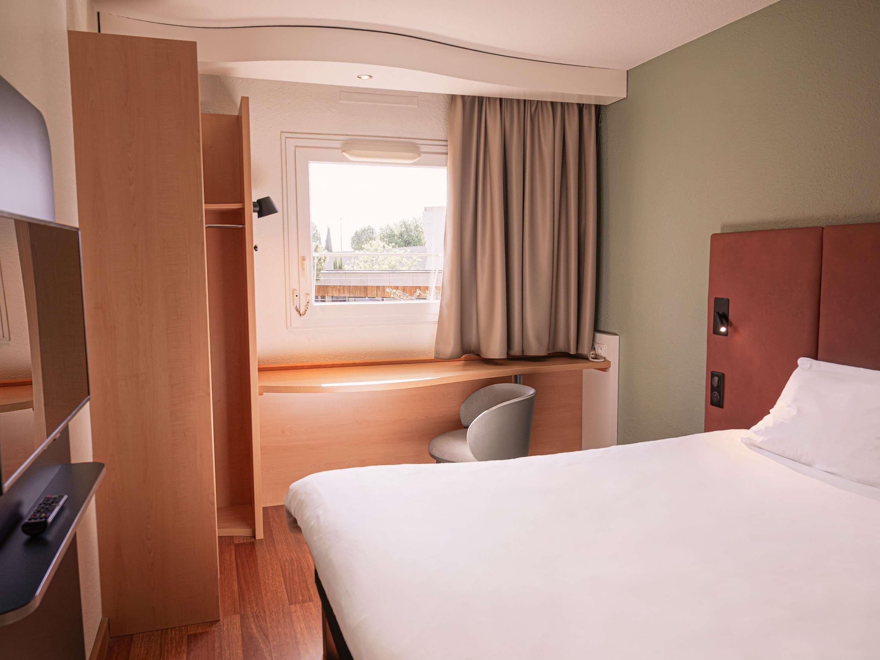 A hotel room with a bed, a desk with a chair, a TV, and a window with curtains.