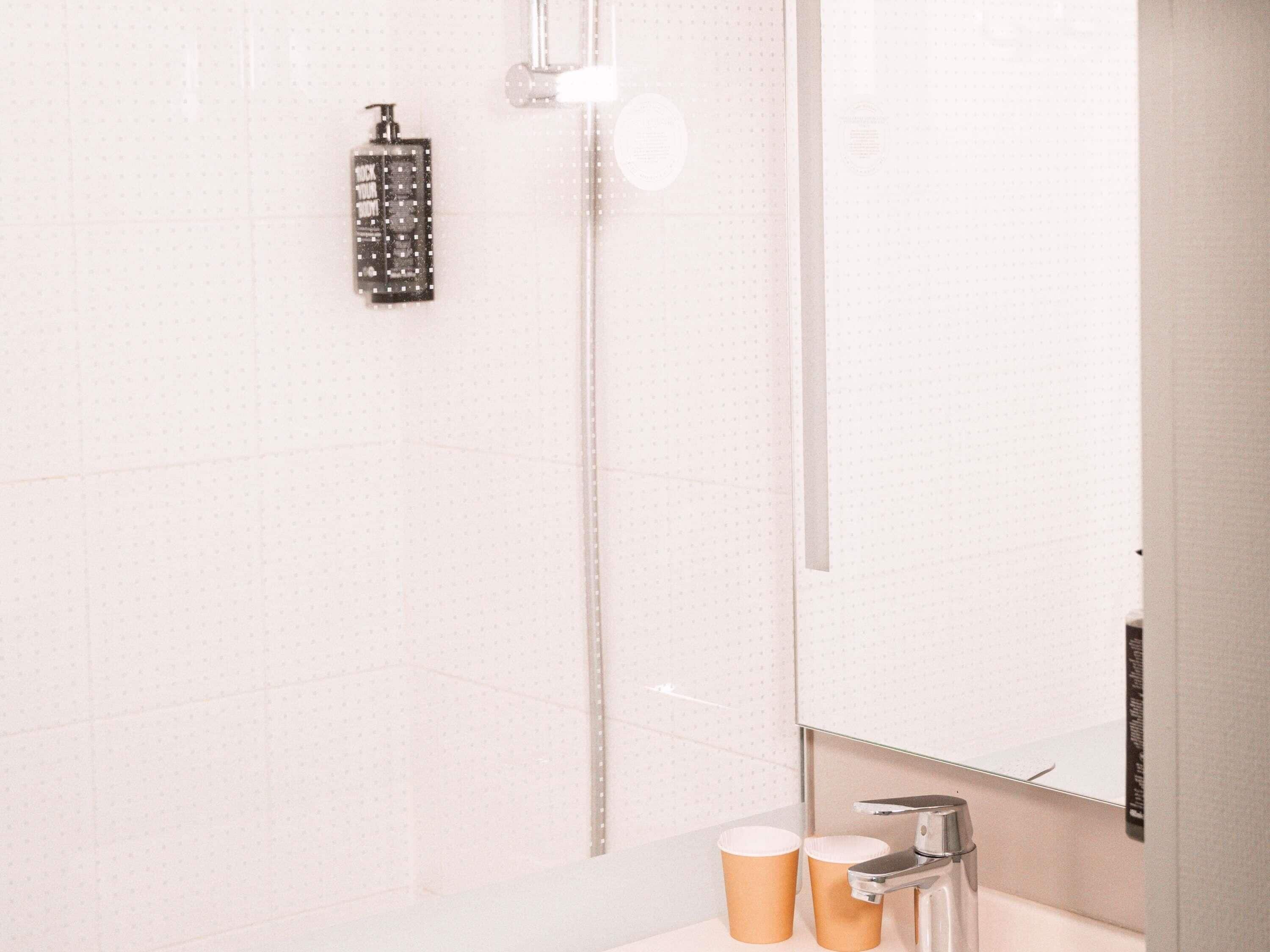 A bathroom with a shower, a mirror, and two paper cups on the counter.