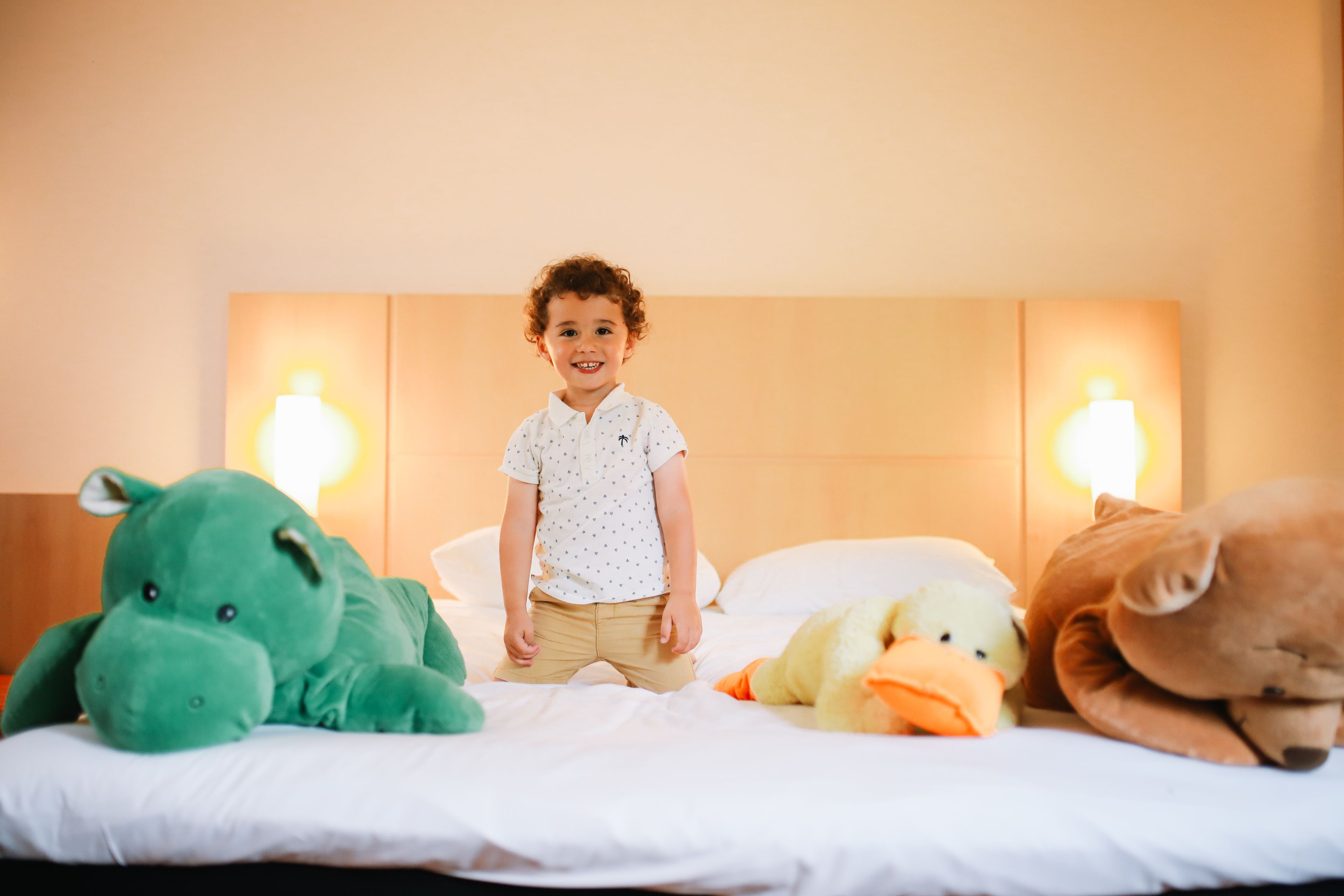 A child standing on a bed with large stuffed animals, a white pillow, and two wall-mounted lamps.