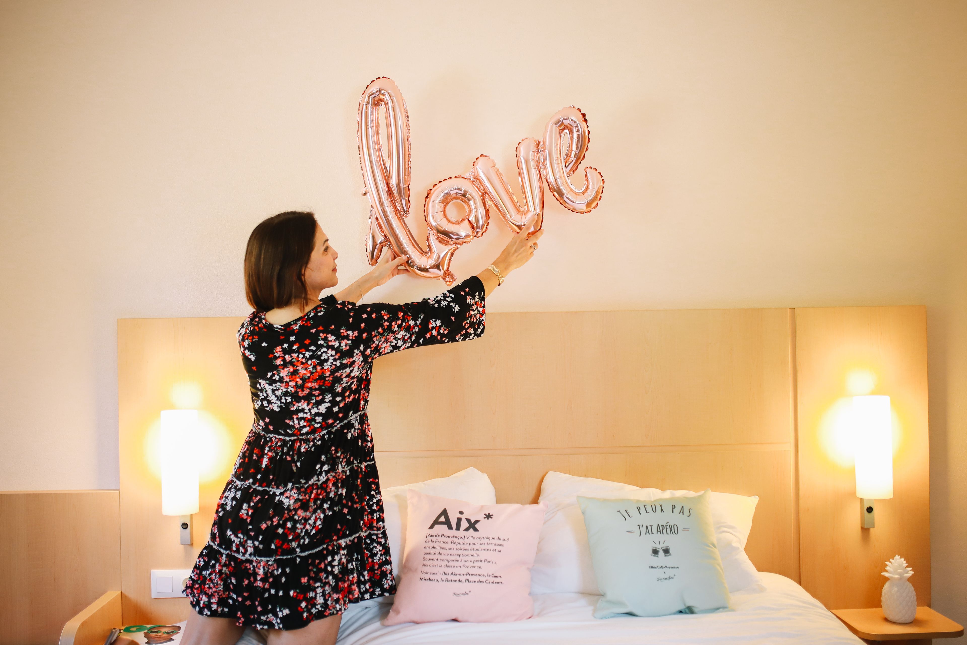 A woman in a floral dress hanging a balloon shaped like the word "love" in a hotel room with a wooden headboard, two bedside lamps, and decorative pillows.