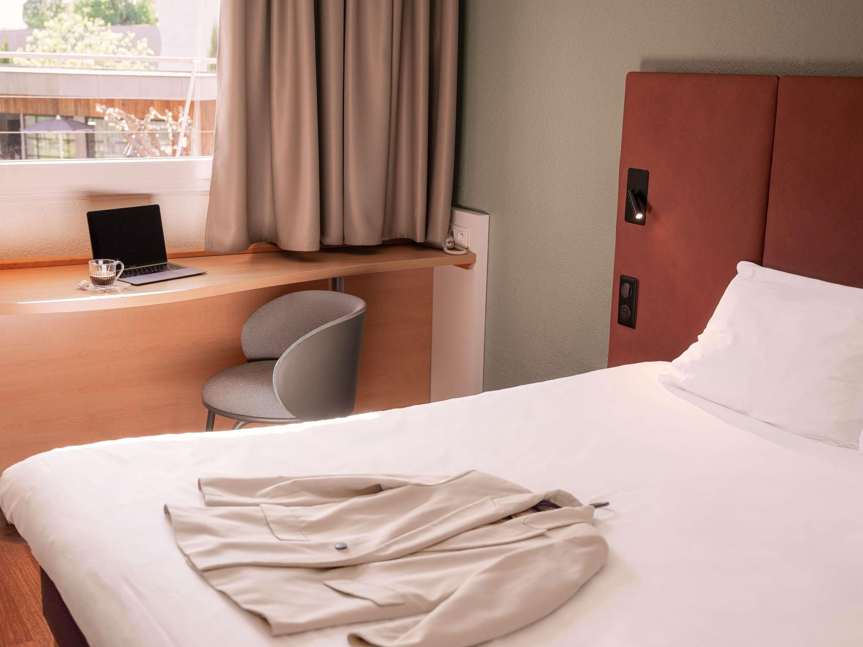 A hotel room with a bed, a desk with a laptop, a chair, and a window with curtains.