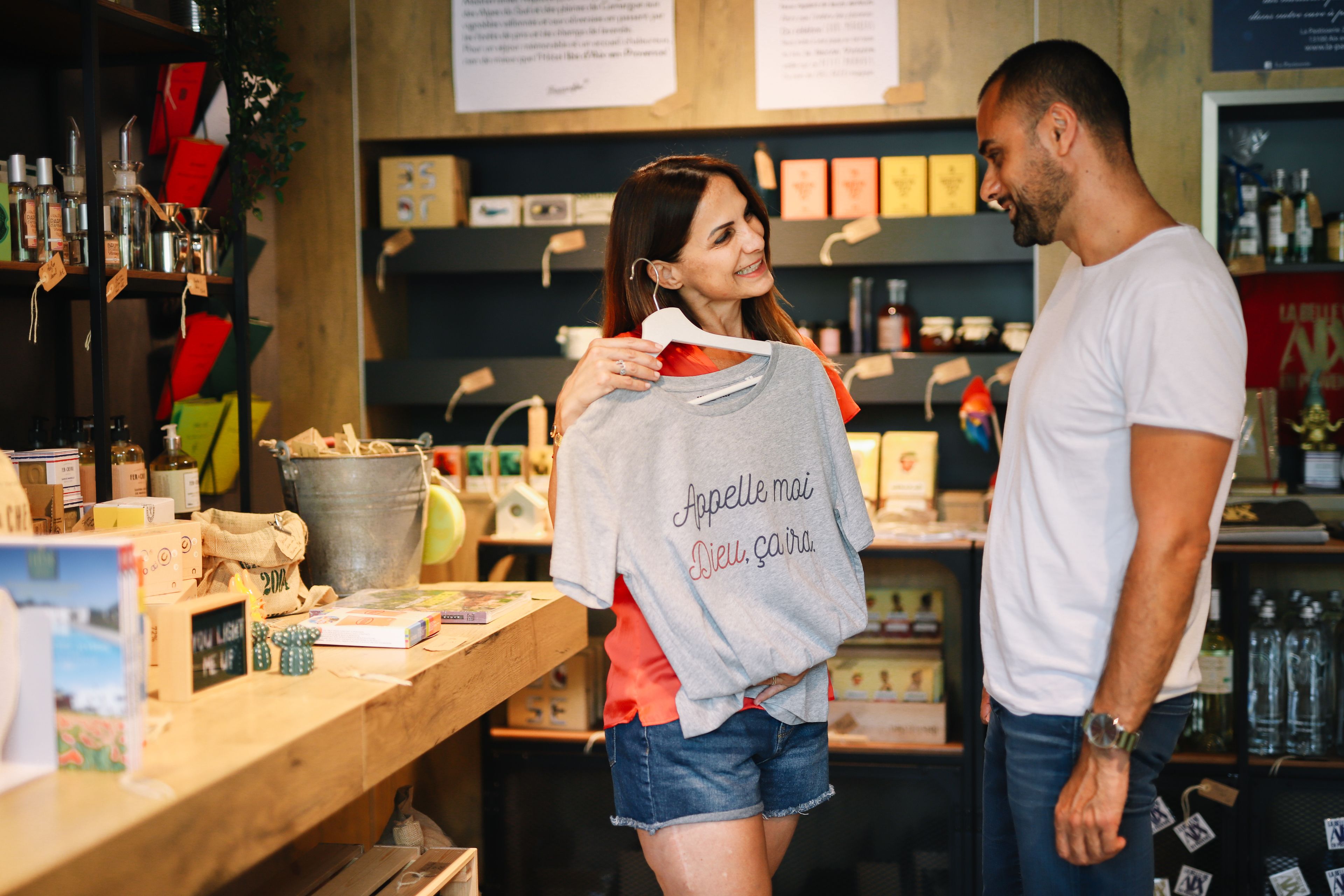 A person holding a grey t-shirt with text, standing in a store with shelves of products and a wooden counter.
