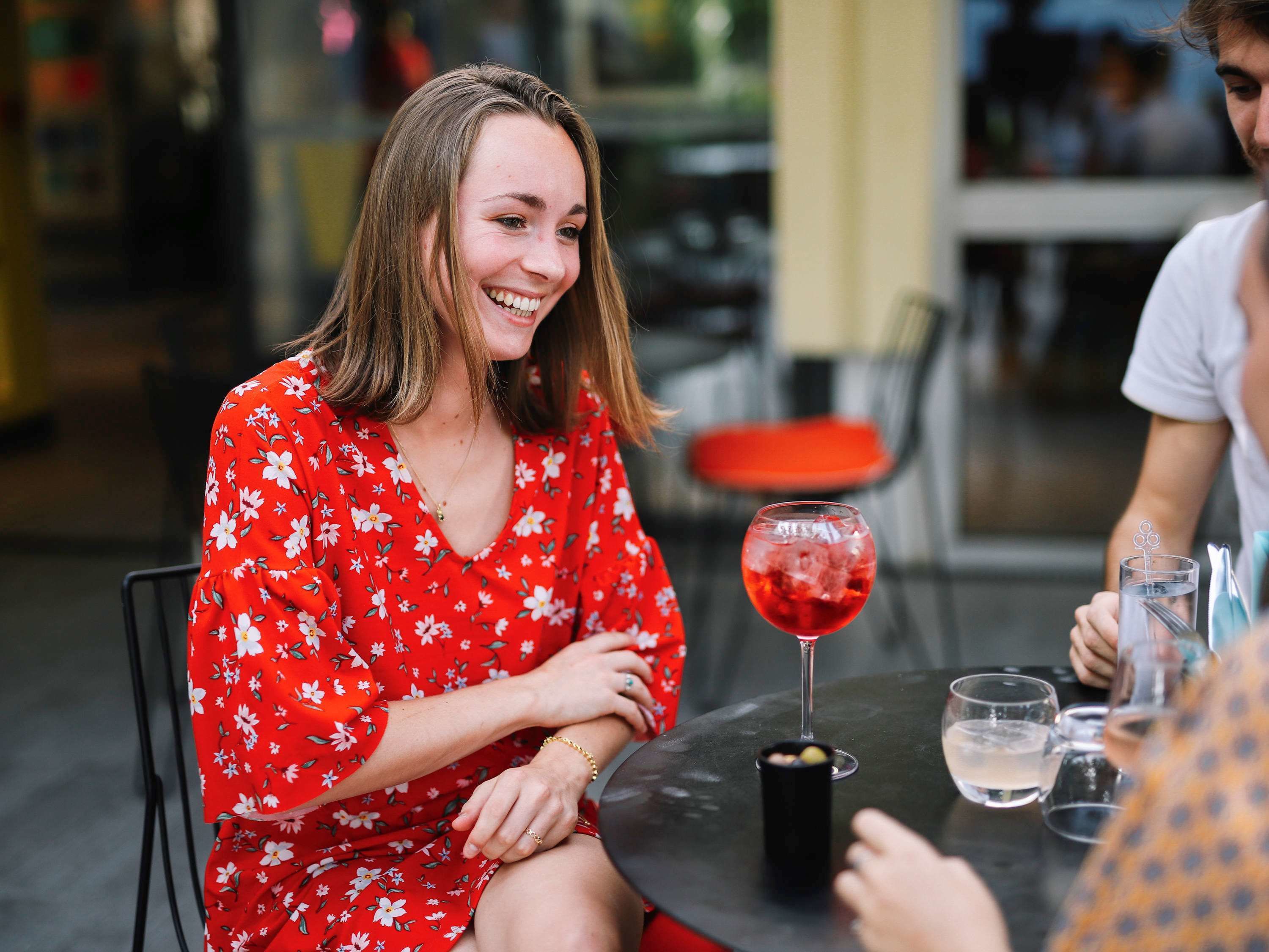 A person in a red floral dress sitting at a table with a glass of red wine and a black cup.