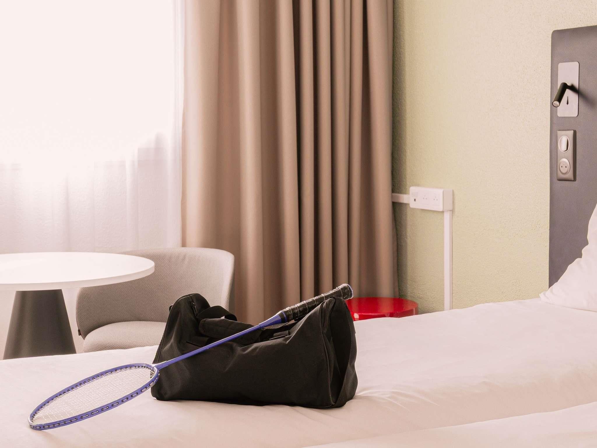 Ein Hotelzimmer mit einem Bett, einer schwarzen Tasche, einem Tennisracket und einem roten Gegenstand auf dem Boden.