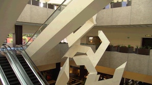 Vista del interior de un edificio con una escalera mecánica y una gran escultura geométrica.