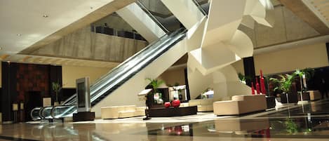 A modern lobby with a central staircase, seating areas, and a glass elevator.