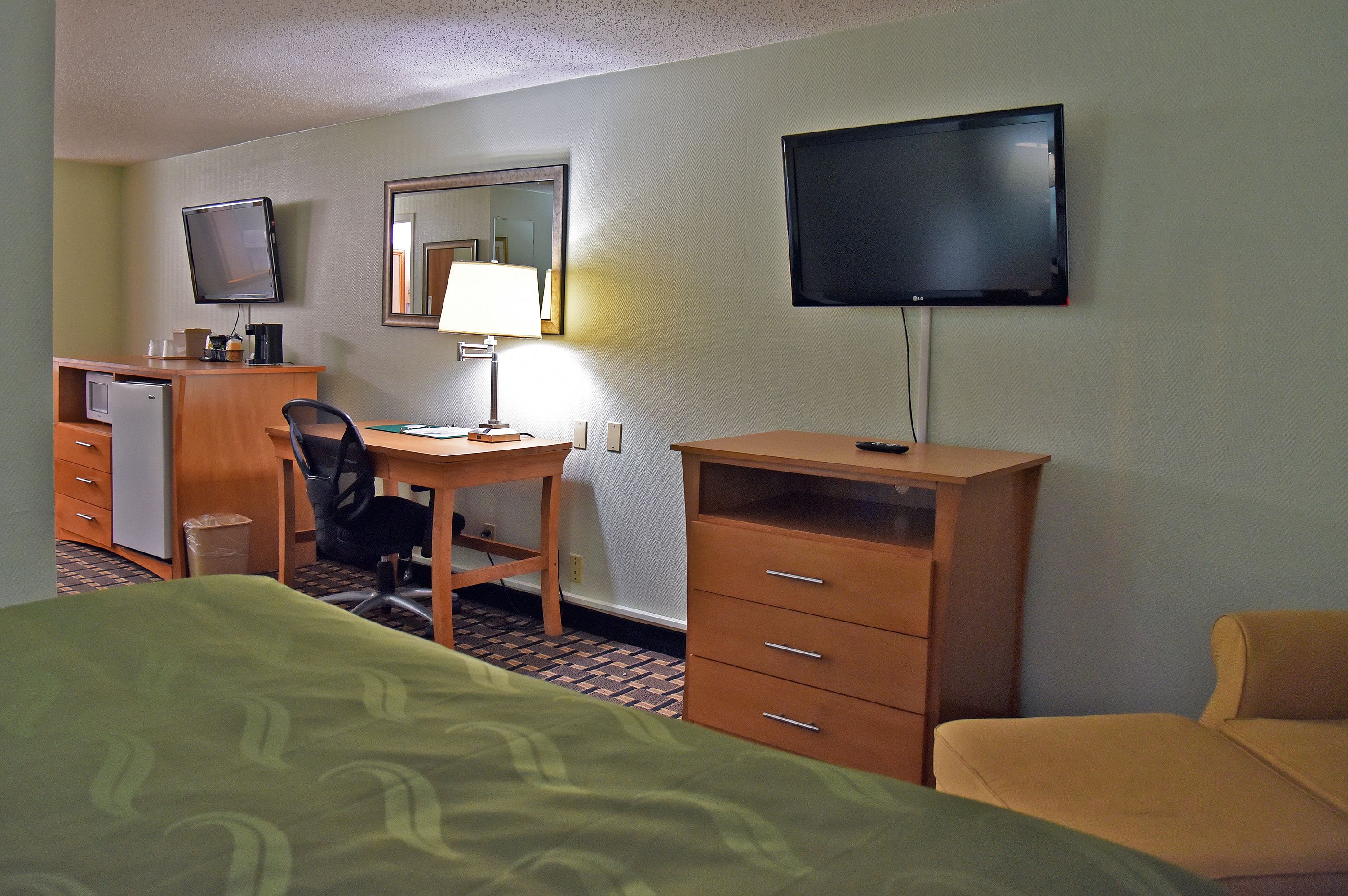 A hotel room with a bed, a desk with a chair, a television, a mirror, and a dresser.