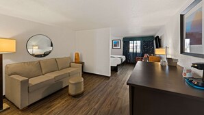 A hotel room with a sofa, ottoman, round mirror, and a bed with a dark headboard. - Quality Inn & Suites Council Bluffs - Omaha East (Council Bluffs)