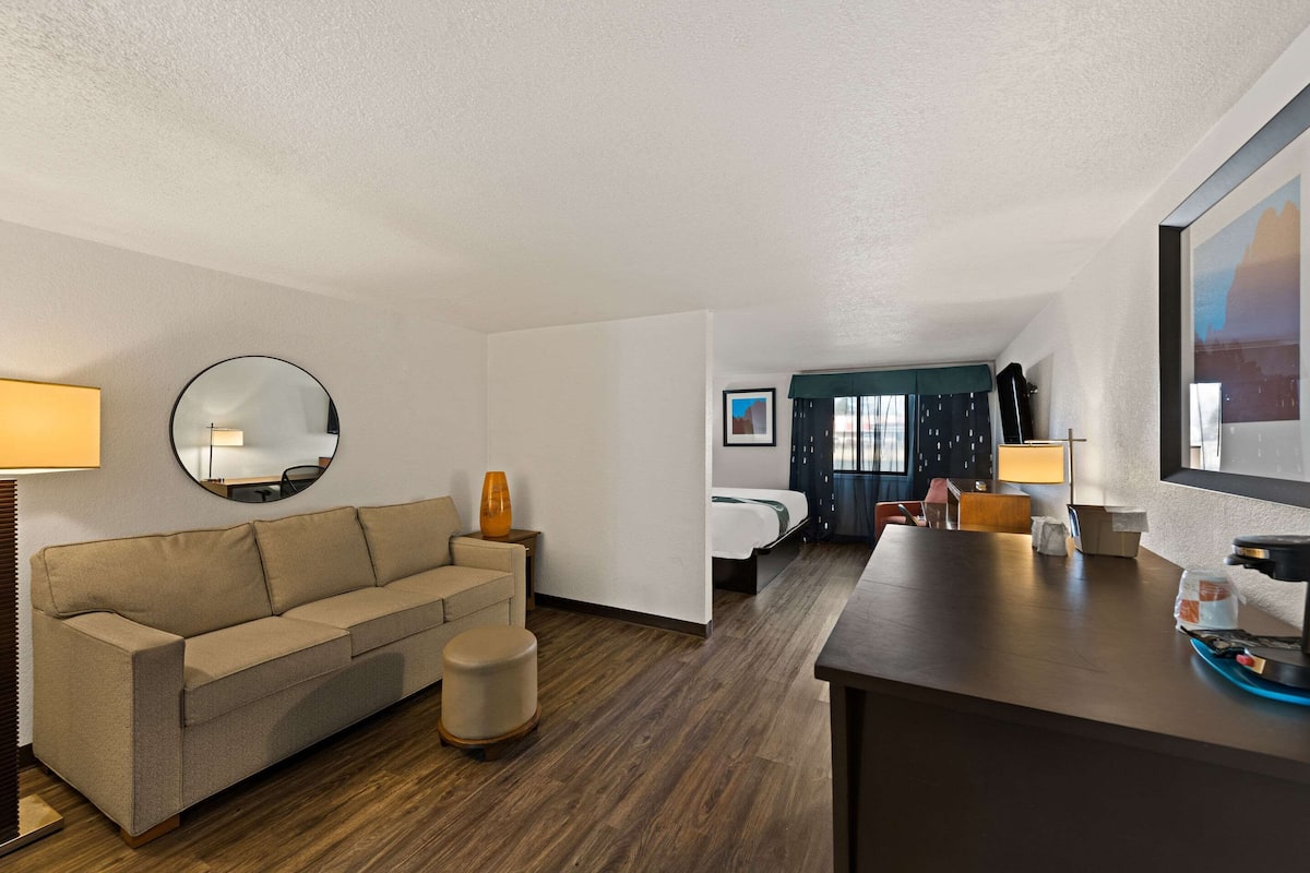 A hotel room with a sofa, ottoman, round mirror, and a bed with a dark headboard.