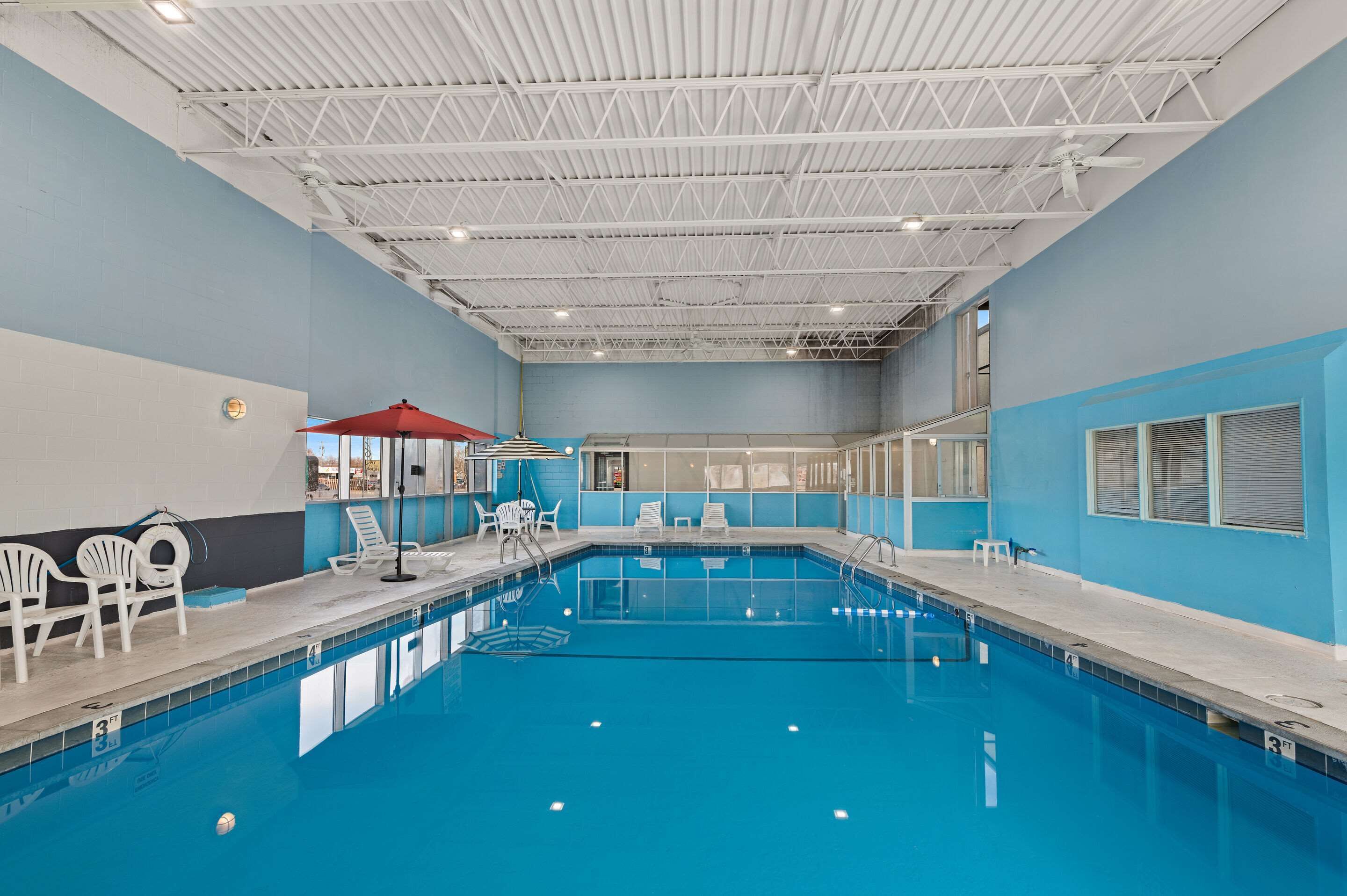 Une piscine intérieure à l’eau claire, avec des chaises longues blanches et un parasol rouge.