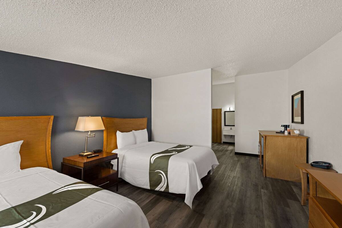 A hotel room with two beds, a desk, a chair, a lamp, and a mirror.