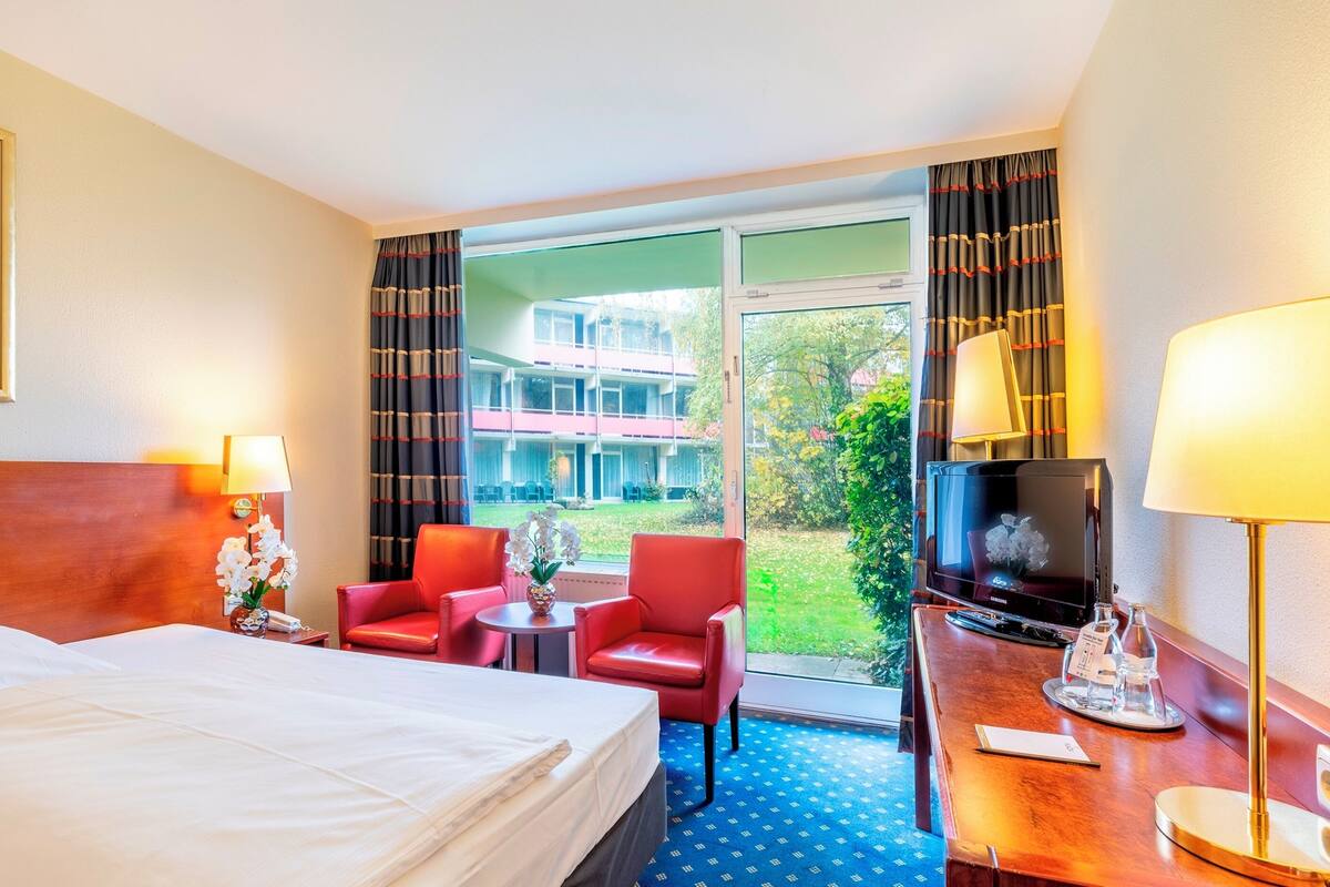A hotel room with a bed, two red chairs, a small table with a vase, a TV, and a desk with a lamp.