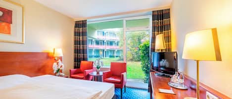 Habitación de hotel con una cama, dos sillas rojas, una mesita con un florero, un televisor y un escritorio con una lámpara.