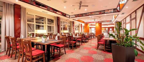 Ein Hotel-Speisesaal mit Holztischen, rot gepolsterten Stühlen, großen Fenstern und einem Deckenventilator.