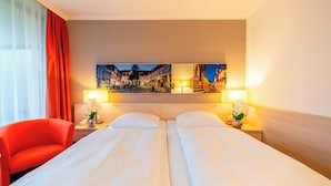 Habitación de hotel con dos camas, una silla roja, una mesita de noche con lámpara y un cuadro de un pueblo europeo en la pared.