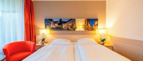 Une chambre d’hôtel avec deux lits, une chaise rouge, une table de chevet avec une lampe et un tableau représentant une ville européenne accroché au mur.