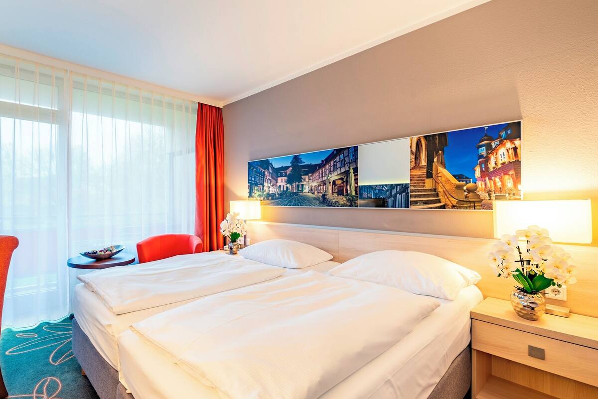 A hotel room with a bed, a desk with a laptop, a TV, and a chair. There is a window with curtains, a nightstand with a lamp, and a painting on the wall.