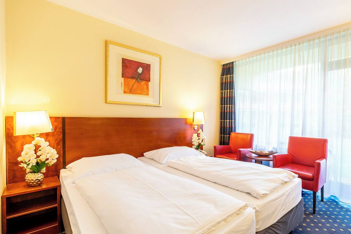 A hotel room with two beds, a red chair, a small table, and a painting on the wall.